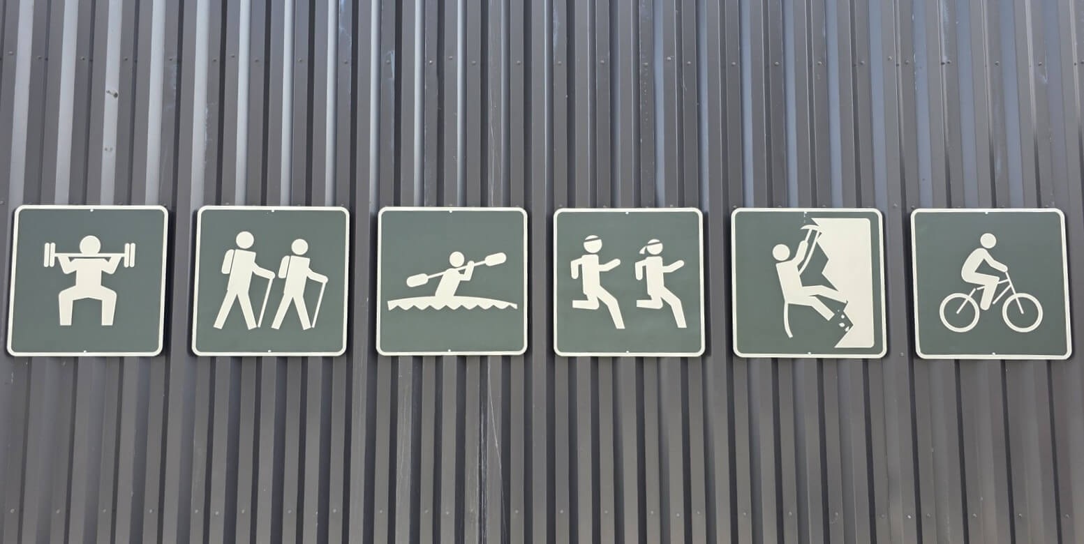Six square signs mounted on a corrugated metal wall, each showing a white pictogram of a different activity: weightlifting, hiking, kayaking, running, rock climbing, and cycling — arranged left to right as a visual catalog of outdoor and fitness pursuits.
