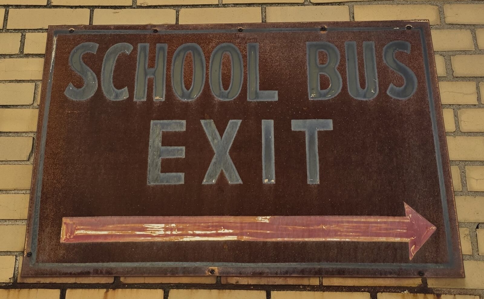 A weathered metal sign mounted on a yellow brick wall reads *School Bus Exit* with a right‑pointing arrow beneath the text.