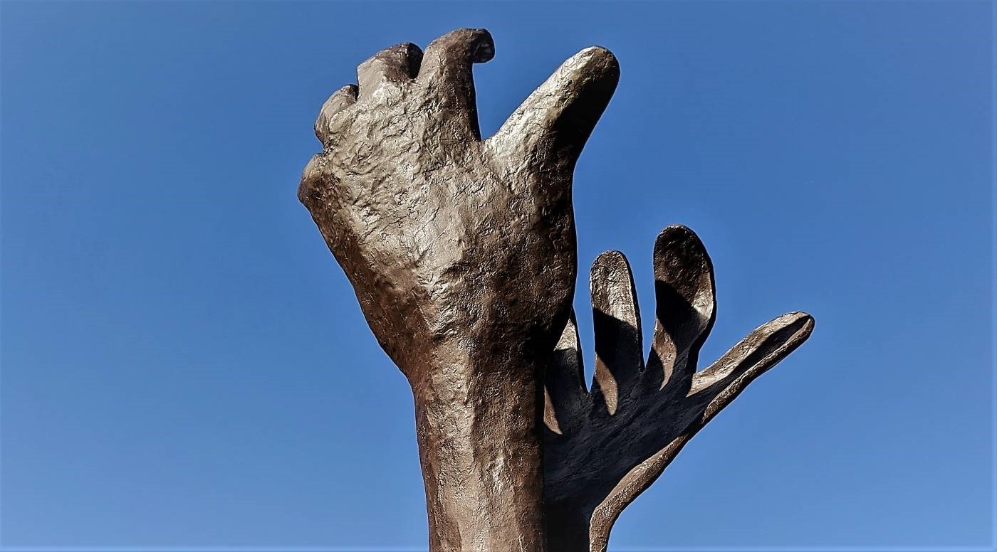 Sculpted brown metalic hands reach up into a blue sky.