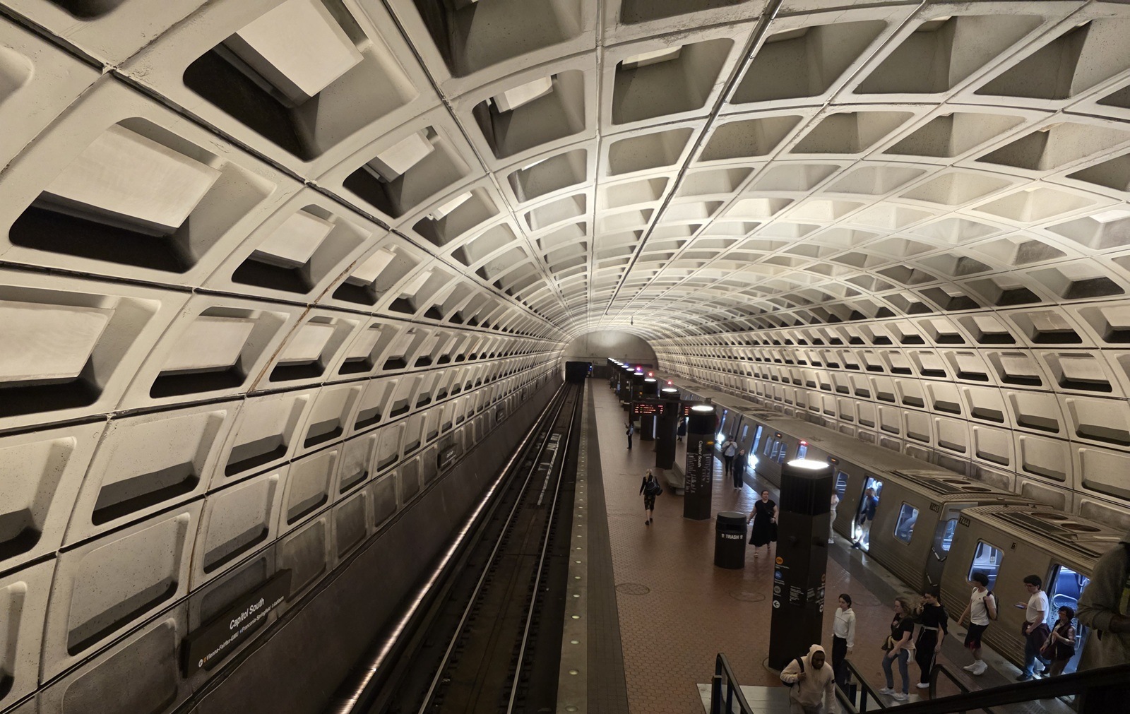 Wide-angle view of the vaulted concrete ceiling and platform of the Capitol South Washington Metro station, with a train stopped on the right side and passengers boarding and exiting. The scene features the station’s iconic Brutalist coffered design and subdued lighting.