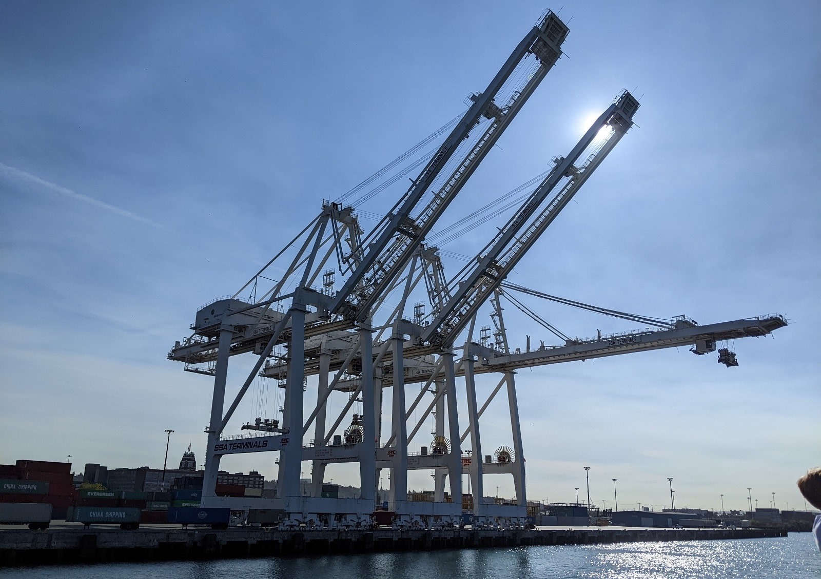 Three massive white cranes on a water front contrasted against a blue sky. The sun is position behind the middle crane.