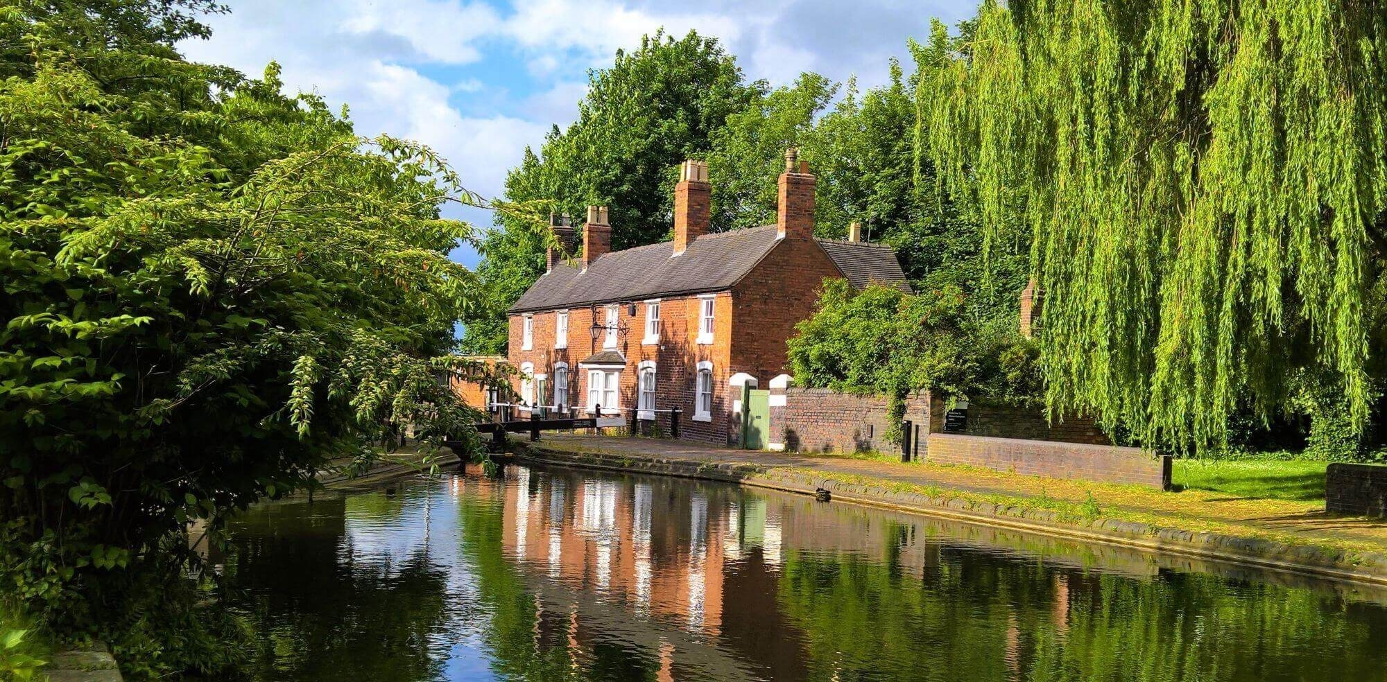 A red-brick canal-side house with white-framed windows and multiple chimneys sits beside a calm waterway, reflecting the building and surrounding greenery. A weeping willow and other lush trees frame the scene, while a small lock gate and brick wall hint at the canal’s navigational use. The sky is partly cloudy, adding to the peaceful, semi-rural atmosphere.