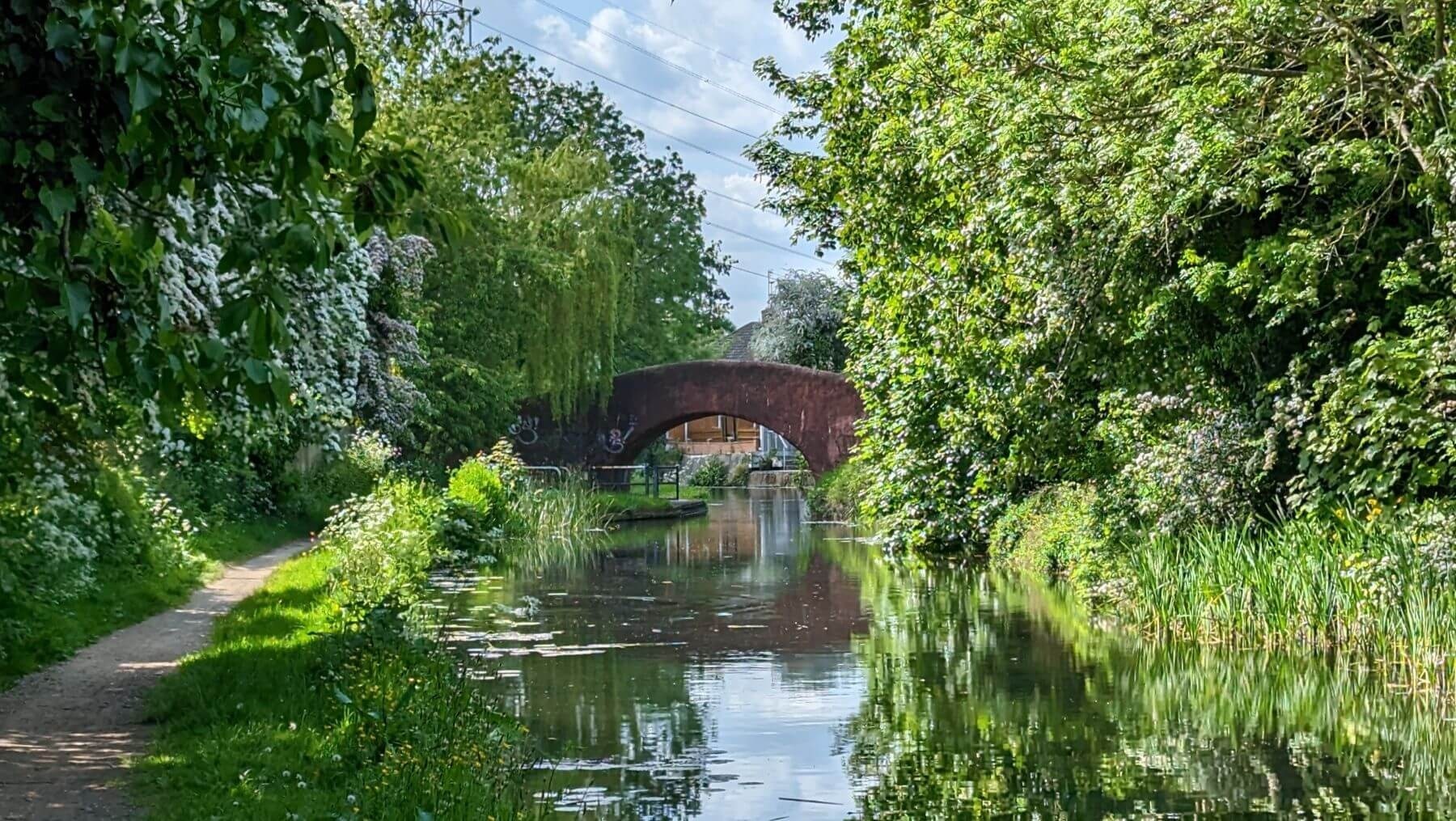 A calm canal scene bordered by lush greenery, with a narrow dirt path running along the left side. Flowering bushes and tall trees reflect in the still water. In the background, a red brick arched bridge spans the canal, partially framing a distant building. Overhead, power lines stretch across a partly cloudy sky, adding a subtle contrast to the peaceful, rural setting.