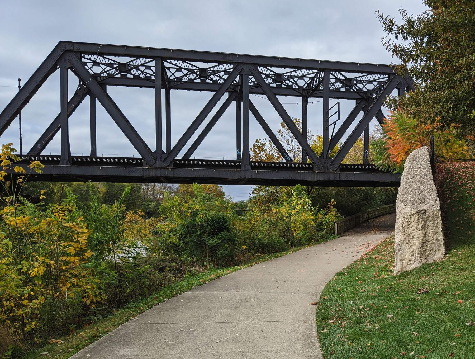 Paved path curving beneath a steel railway bridge, beside a grassy bank and trees with autumn foliage.